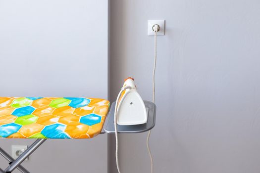 Electric iron plugged into an outlet on an ironing board against a gray wall. Ironing clothes.
