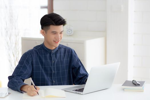 Young asian business man writing on notebook for planning working and using laptop computer on desk at home, notes about finance, male study and learning, business and communication concept.