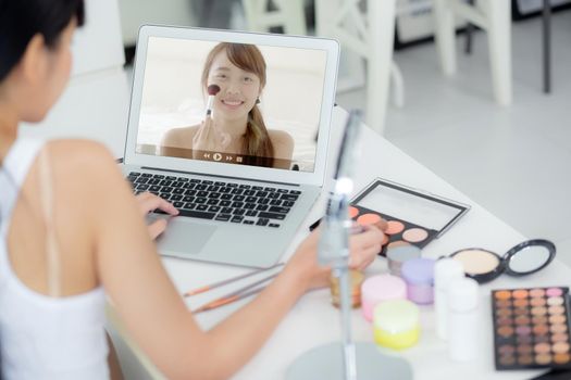 Beauty of young asian woman with learning makeup with brush on cheek on laptop computer with tutorial course online, female teaching and explain make up with cosmetic on streaming social media.