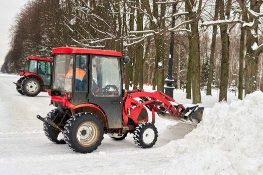 Red tractor with a large bucket removes snow in a city park. Close up