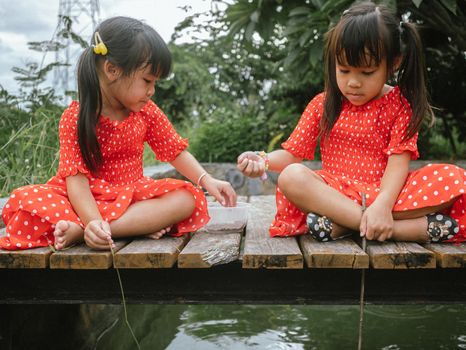 Two little sibling girls in red dress role playing fishing sitting on a wooden bridge by the pond. Childhood happiness.
