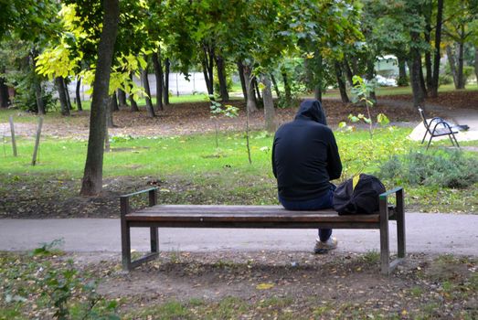 Young man in black jacket sitting alone on bench between trees at beautiful day at park. Autumn. Thinking about life. Spending time alone in nature. Peaceful atmosphere. Back view. High quality photo