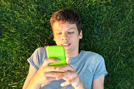 Guy teenager lying on grass with smartphone. Male in gray T-shirt with phone in his hands, top view, green lawn background. Youth, technology, lifestyle, adolescence concept