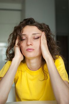 Tired young woman with curly hair and yellow shirt is working from home using her laptop at the kitchen table in her apartment, remote work, freelance, burnout syndrome.