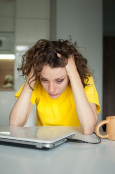 Tired young woman with curly hair and yellow shirt is working from home using her laptop at the kitchen table in her apartment, remote work, freelance, burnout syndrome.