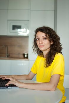 Young woman with curly hair and yellow shirt is working from home using her laptop at the kitchen table in her apartment, remote work, freelance.