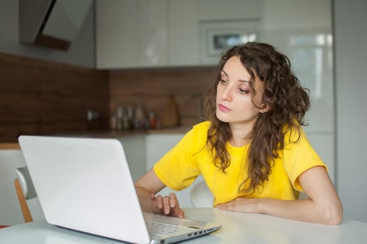Young woman with curly hair and yellow shirt is working from home using her laptop at the kitchen table in her apartment, remote work, freelance.