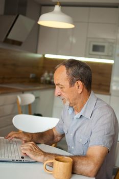 Trendy mature man is working from home with laptop sitting at the table in his kitchen, happy retirement, home-office concepts.