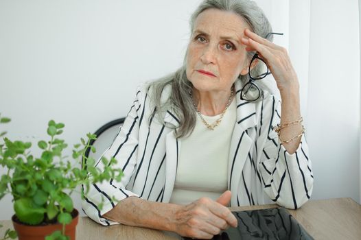 Tired senior grey haired businesswoman in striped jacket with eyeglasses is working in her office sitting at the desk and feeling bad due to menopause, menopause relief concept.