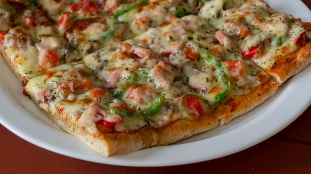 Close up on a white plate with a square pizza with ham, green pepper, tomato, olives and cheese cut into rectangular pieces on wooden table