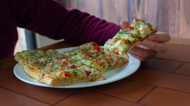 Woman's hand taking a rectangular piece of square pizza with ham, green pepper, tomato, olives and cheese from a white plate on a wooden table