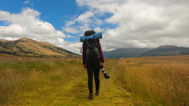 Beautiful Hispanic female explorer seen from behind with backpack walking with a camera in hand in the middle of a sown field on a cloudy morning