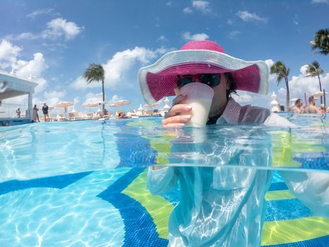 Woman wearing hat staning in pool drinking cocktail. Concept of happy vacation
