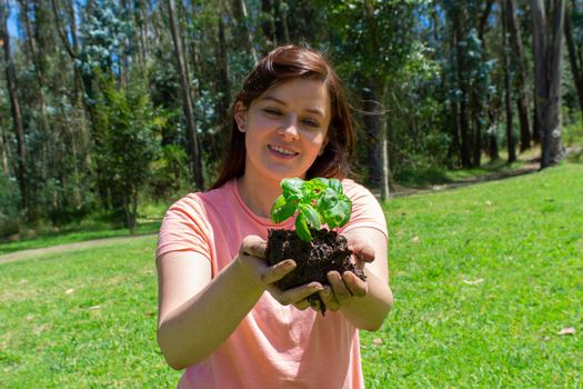 Beautiful young Hispanic woman holding a small plant in her field hands before being planted in a green field surrounded by trees during the morning