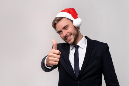 Young handsome caucasian guy in business suit and Santa hats on white background in studio smilie and showing thumbs up two hands. Close up portrait business person with Christmas mood Holiday banner