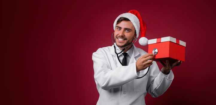 Young handsome doctor in white uniforme and Santa Claus hat standing in studio on red background smile Holding red gift box in hand Portrait medical student Christmas New Year Holiday Medicine concept