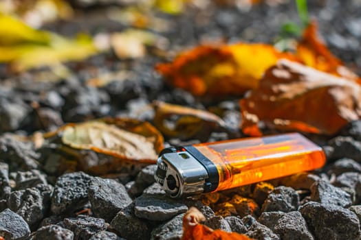 A bright orange lighter lies on the stones among fallen leaves, thrown onto the road by someone
