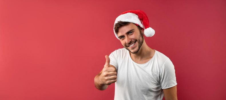Young handsome caucasian guy in a white t-shirt and Santa hats stands on red background in studio and showing thumbs up. Close up portrait european guy with Christmas mood. Holiday banner.
