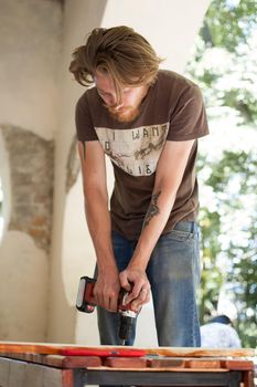 13.08.2021 - Ukraine, Goshcha, voluntary event, skilled young male worker is using power screwdriver drilling during construction wooden bench, do it yourself.