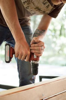 13.08.2021 - Ukraine, Goshcha, voluntary event, skilled young male worker is using power screwdriver drilling during construction wooden bench, do it yourself.