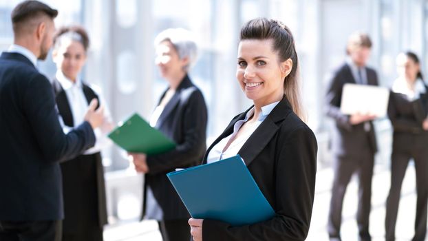 Beautiful young business woman in formal suit standing in front of colleagues holding document folder in business building