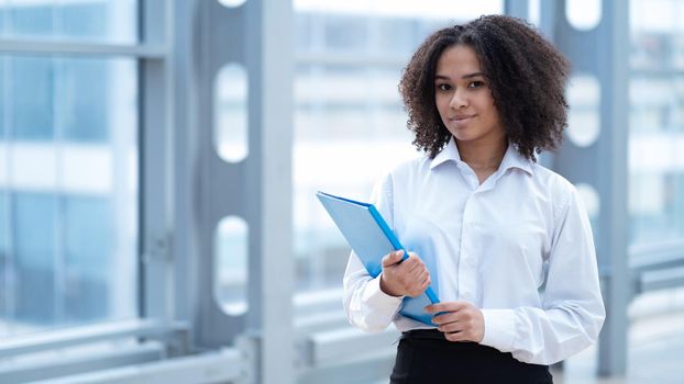 Happy african business woman holding folder of documents standing near window in office