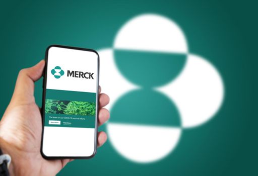 New York, USA, October 2021: A hand holding a phone with the Merck pharmaceutical company app on the screen and Merck logo blurred on a green background.