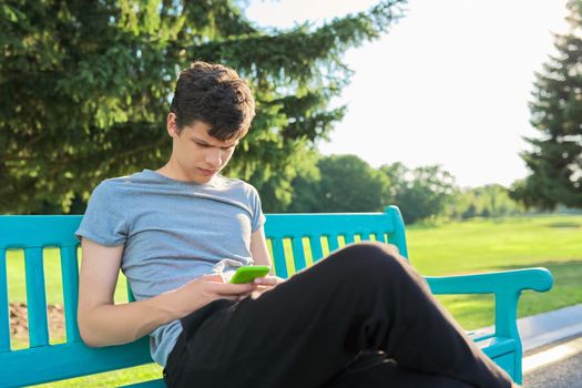 Serious guy teenager using smartphone, having rest, sitting on bench in park. Youth, technology, lifestyle, adolescence concept