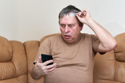 Senior citizen sitting on the couch understands the gadget. Man took off his glasses and opened his mouth in surprise