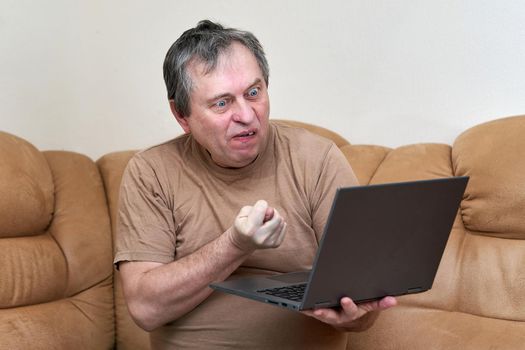 A man at the age of sitting on Devani looks through the news on a laptop. Man shows fig in laptop screen