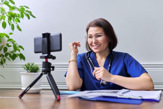 Female doctor with stethoscope consulting online using smartphone on tripod. Talking doctor looking at phone webcam, video meeting with patient. Medicine, technology, lifestyle, profession, people concept