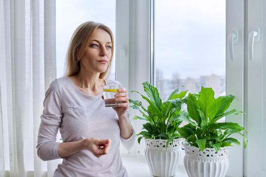 Middle-aged woman drinking lemon water at home near the window, winter autumn season, natural vitamins. Health, nutrition, natural antioxidant, beauty, 40s, mature people, lifestyle concept