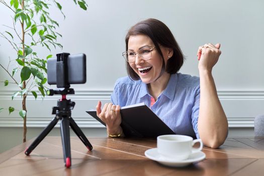 Middle-aged woman teacher, business woman working online using smartphone. Business positive female with notepad on table talking looking in webcam phone on tripod, at home. Technology education work