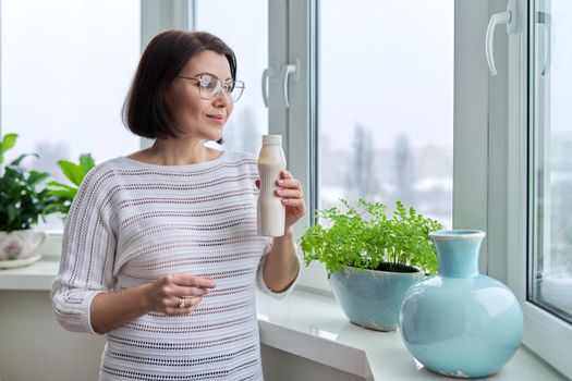 Middle-aged woman drinking milk drink, liquid yogurt in a bottle, at home near the window. Healthy food, lifestyle, winter season, mature age, nutrition, diet concept