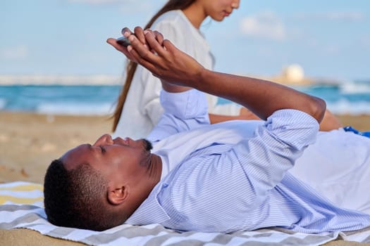 Young beautiful couple having a rest on the beach, African American man with a smartphone in his hands close-up. Lifestyle, technology, leisure, people concept