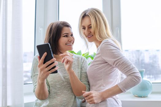 Two middle-aged women friends looking together at smartphone screen. Females at home near window in winter season. Relationship, lifestyle, leisure, friendship, technology, mature people concept