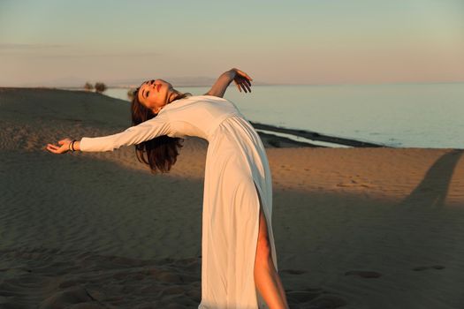 A girl in a fly white dress dances and poses in the sand desert at sunset.