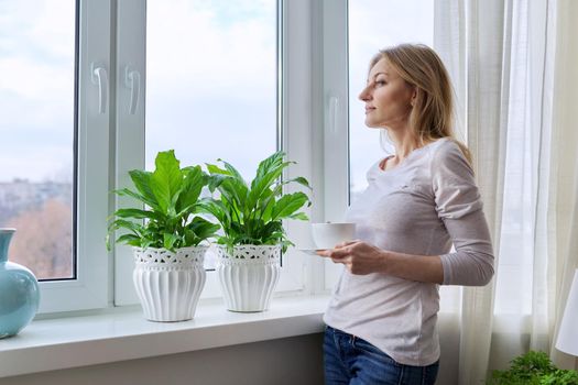 Middle aged beautiful woman with a cup of coffee at home near the window in the winter autumn season, copy space. Beauty, lifestyle, comfort, tranquility, mature age concept