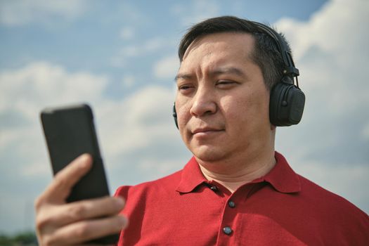 Middle-aged Asian man in headphones outdoors listening to music against the background of the sky, mobile phone in his hand