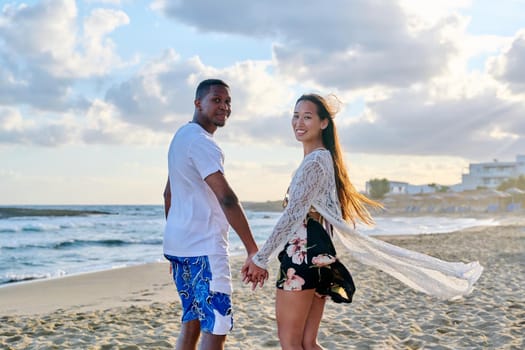 Portrait of beautiful young multiethnic couple holding hands on the seashore. Happy romantic loving african man and asian woman looking in camera. People, love, relationship, vacation, tourism concept