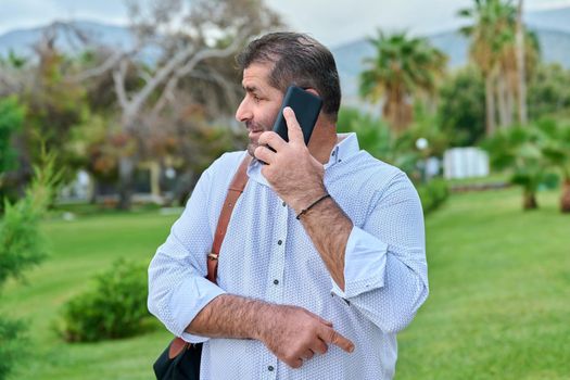 Mature business man talking on the phone outdoors. Serious confident male with backpack, in tropical park. Business trip, freelance, communication, technology, middle aged people concept