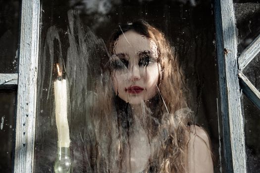 Beautiful sad young girl looking out the window with a candle, good for book cover