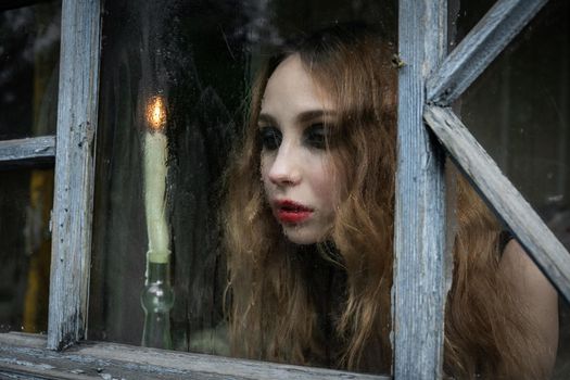 Beautiful sad young girl looking out the window with a candle, good for book cover