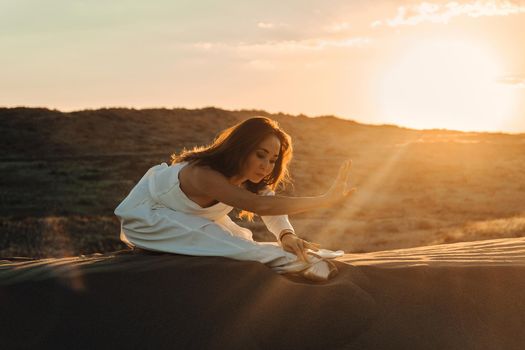 A girl in a fly white dress dances and poses in the sand desert at sunset.