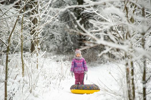 a little girl in winter in purple clothes and an inflatable circle walks on the street in a snow-covered forest.