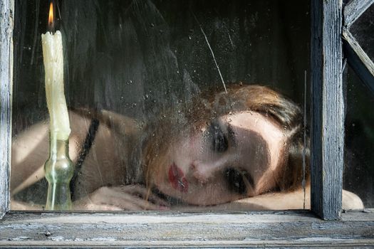 Beautiful sad young girl looking out the window with a candle, good for book cover