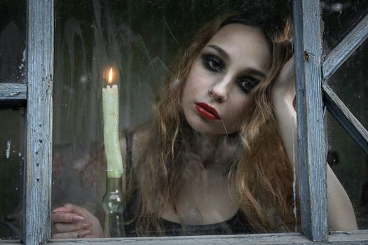 Beautiful sad young girl looking out the window with a candle, good for book cover