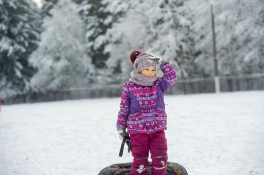 a little girl in winter in purple clothes and an inflatable circle walks on the street in a snow-covered forest.