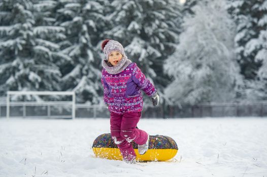 a little girl in winter in purple clothes and an inflatable circle walks on the street in a snow-covered forest.