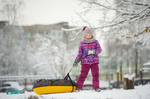 a little girl in winter in purple clothes and an inflatable circle walks on the street in a snow-covered forest.